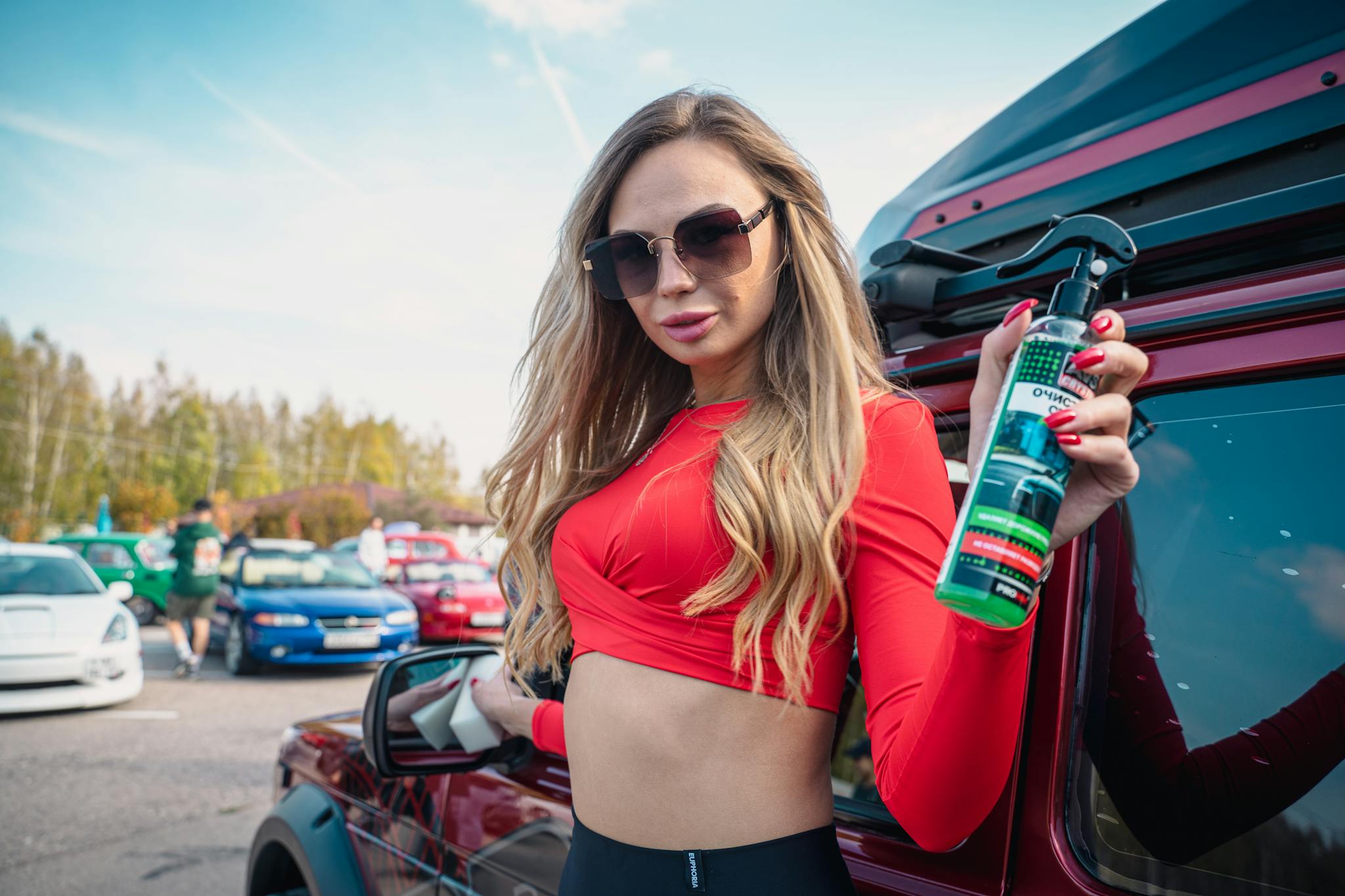 Young woman posing outdoors with car cleaner product, wearing sunglasses and a red crop top.